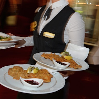 Eine Servicekraft trägt drei Teller mit Schnitzeln durch ein Restaurant.