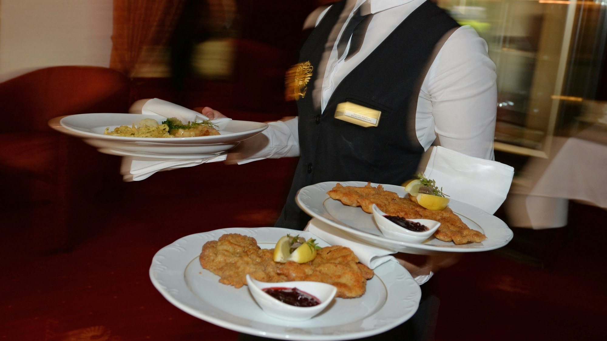 Eine Servicekraft trägt drei Teller mit Schnitzeln durch ein Restaurant.