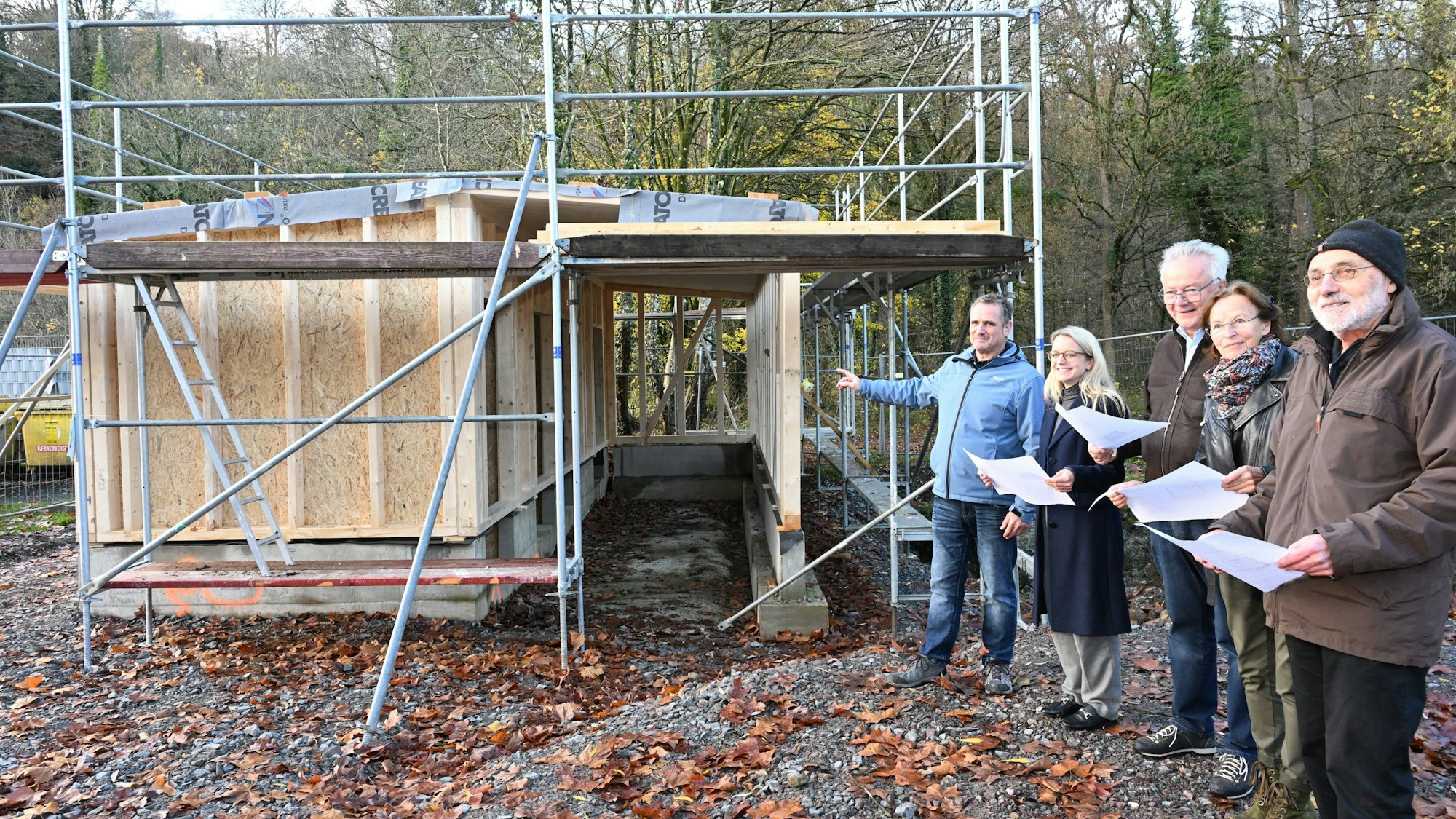 Fünf Menschen stehen an einem kleinen Rohbau und halten Baupläne in der Hand.