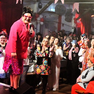 Lieselotte Lotterlappen steht mit Mikrofon in der Hand vor den jubelnden Jecken.