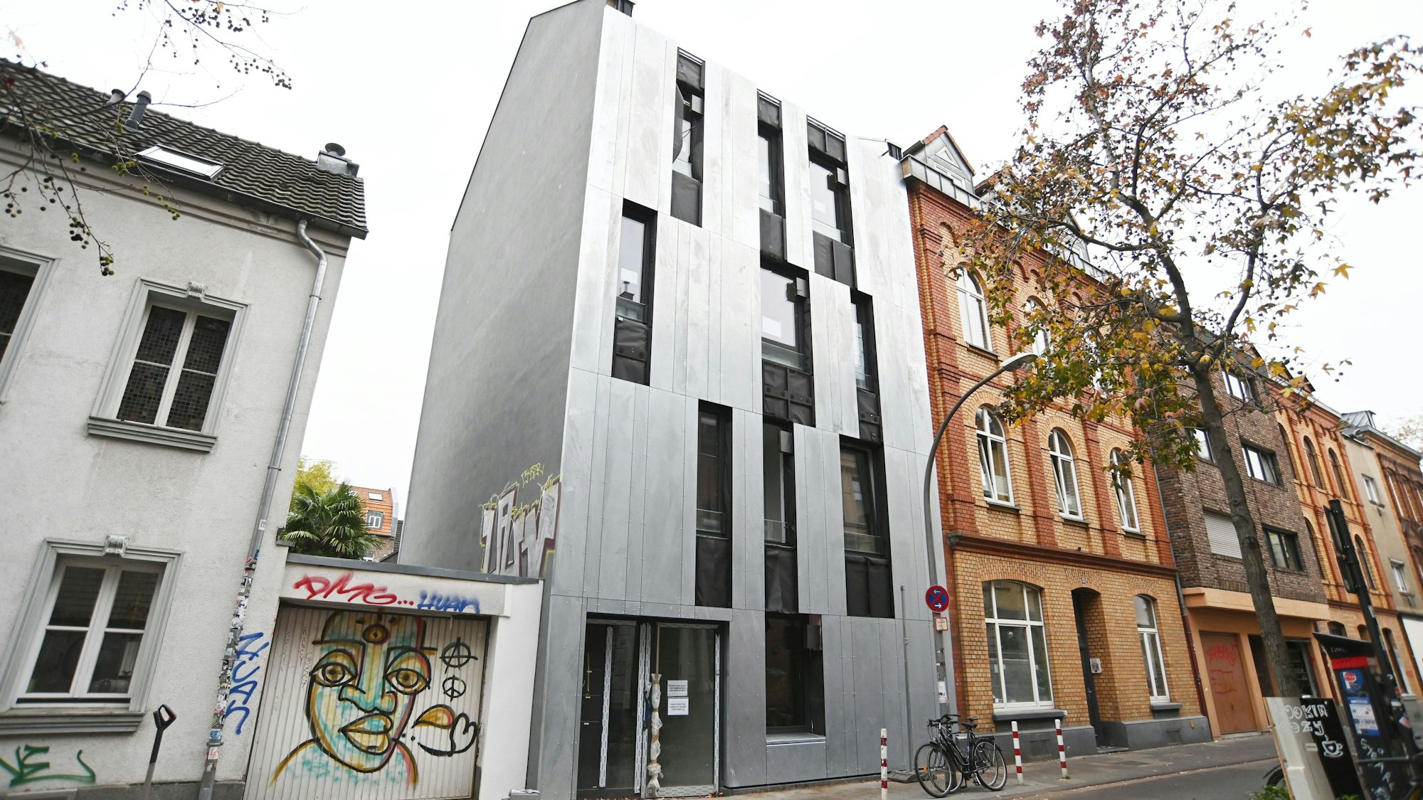 An einer Straße sticht ein Neubau heraus, seine Fassade ist silberfarben.