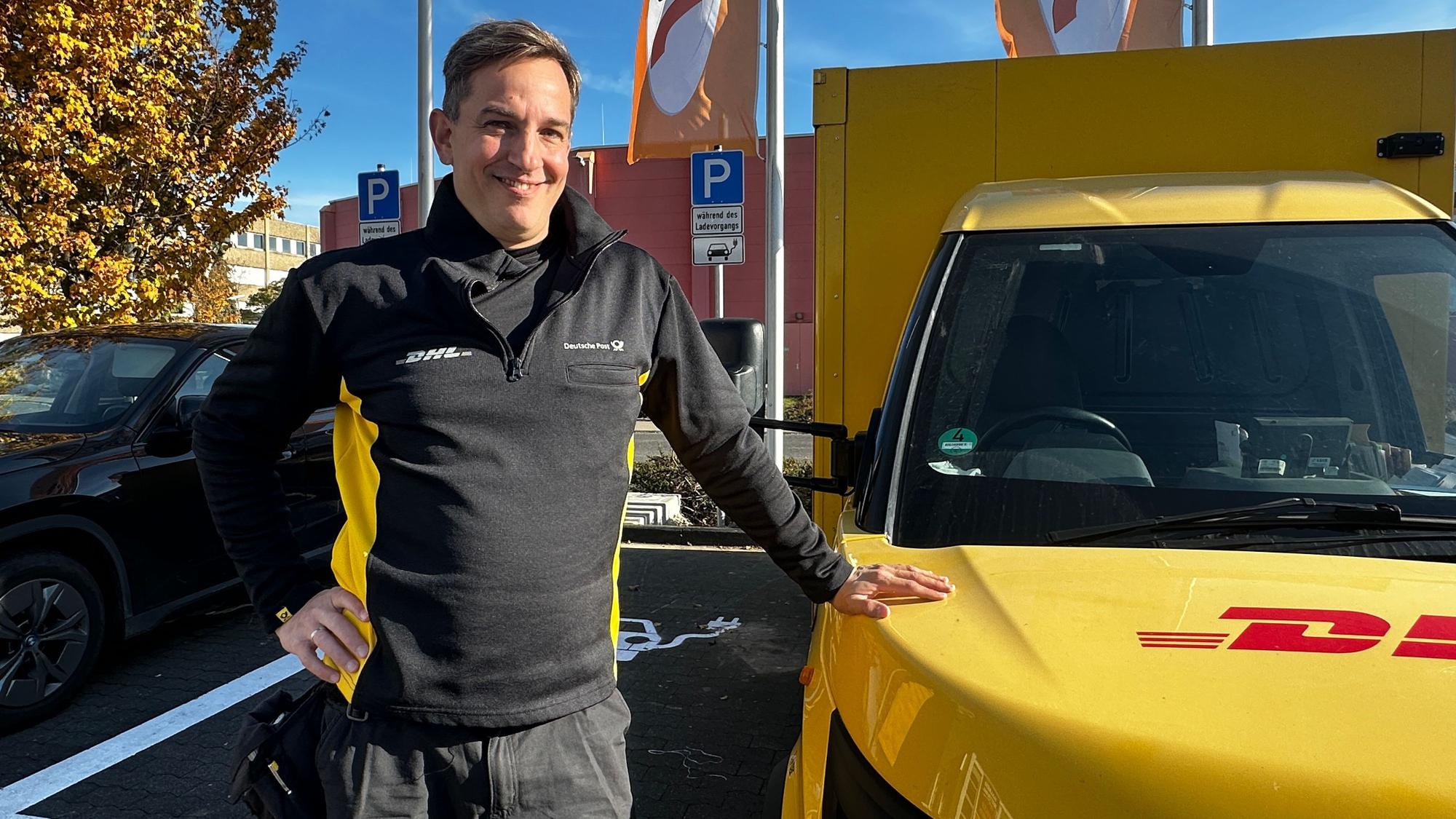 Ein Mann in Postuniform steht neben einem gelben DHL-Zustell-Transporter.
