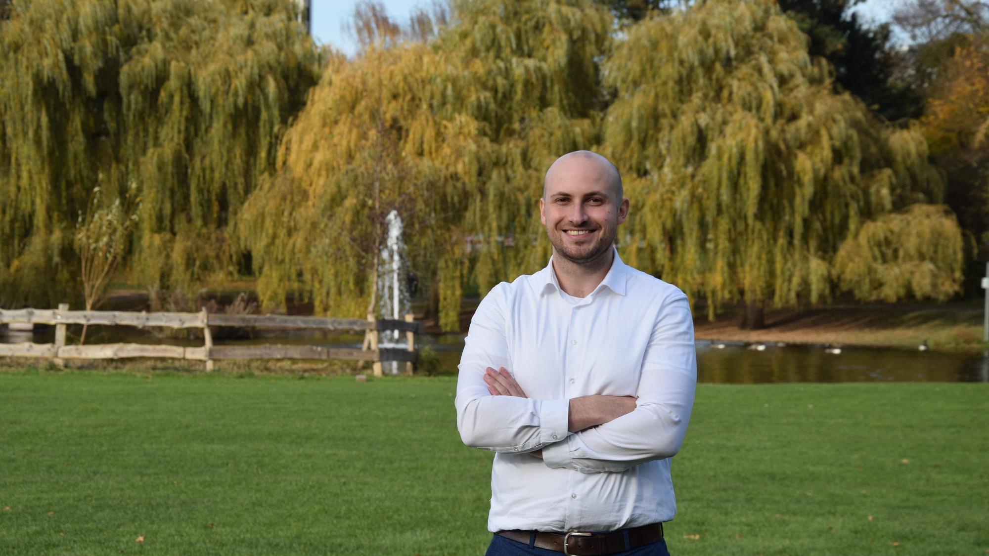 Ein junger Mann mit Glatze und Bart steht mit verschränkten Armen vor einem Teich in einem Park.