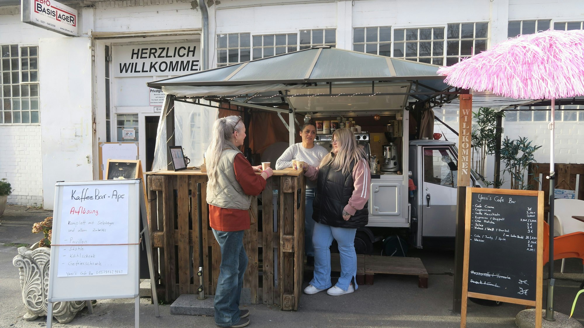 Drei Frauen stehen vor der hölzernen Theke eines Kaffeemobils, das vor einer großen Halle steht.