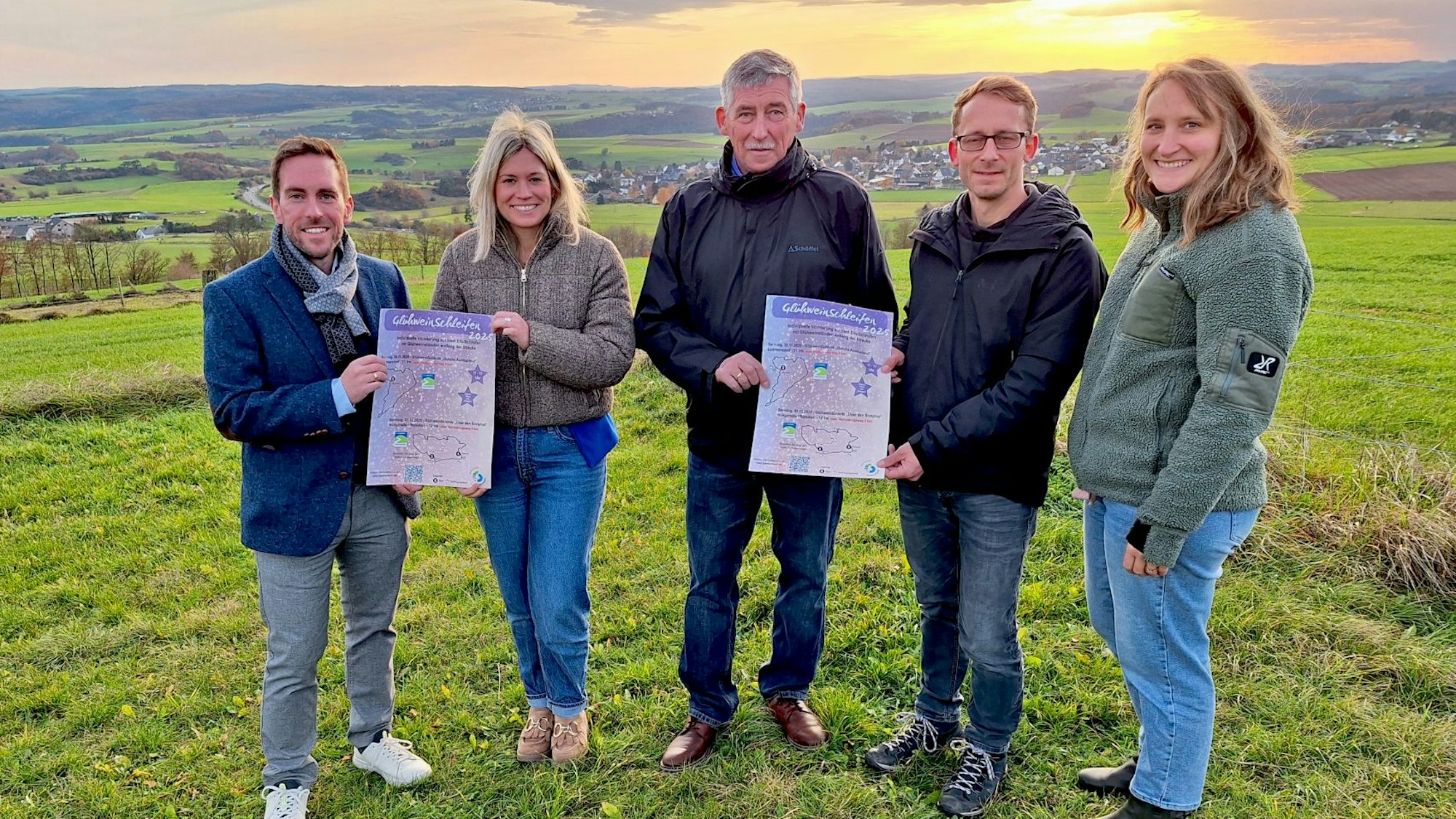 Drei Männer und zwei Frauen stehen bei Sonnenuntergang auf einer Wiese und präsentieren zwei Veranstaltungsplakate.