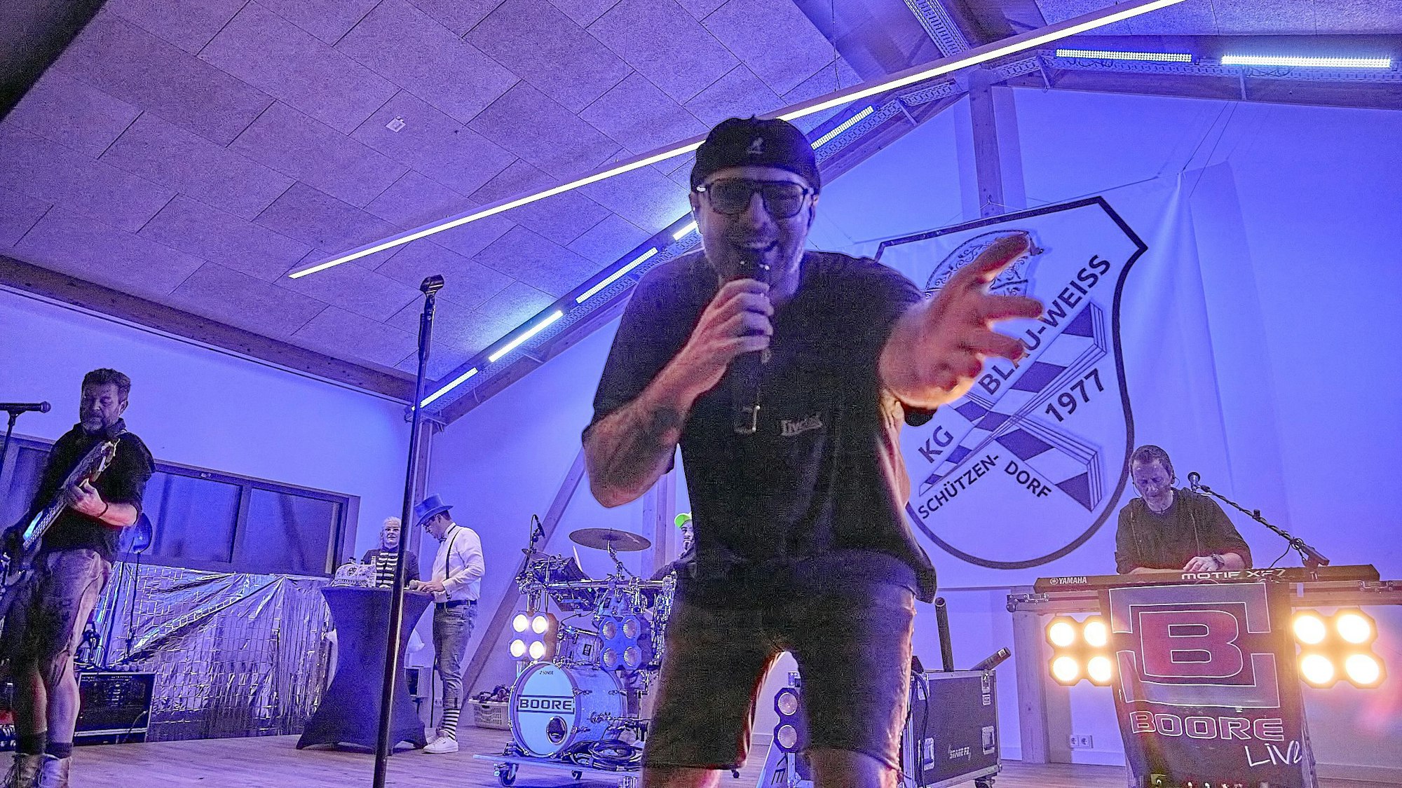 Der Frontmann der Boore beugt sich ins Mikrofon singend in Richtung Kamera, im Hintergrund sieht man die Bandkollegen musizieren.