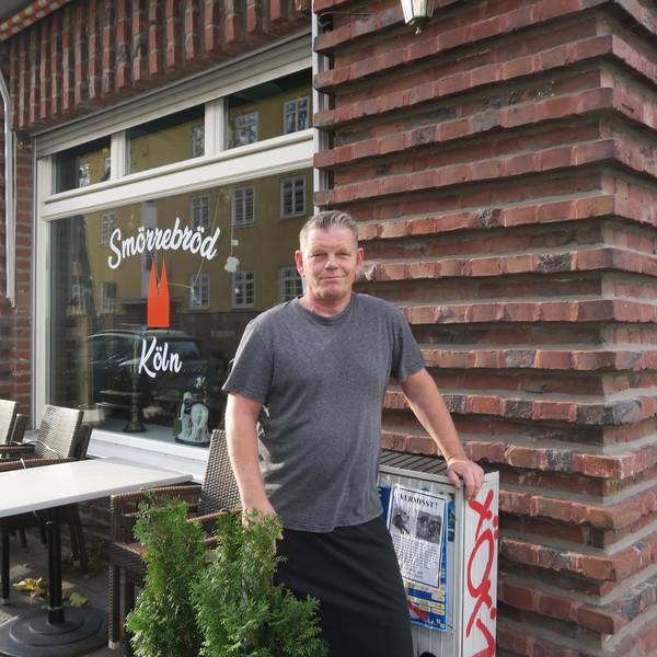 Ein Mann im grauen T-Shirt steht vor einem Lokal. Auch Tische und Stühle stehen vor der Fensterfront des Ladenlokals.