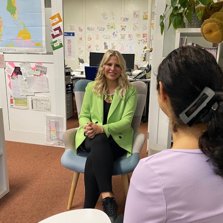 Eine Frau mit dunklen Haaren ist von hinten zu sehen. Sie redet mit einer blonden Frau, die gegenüber sitzt.