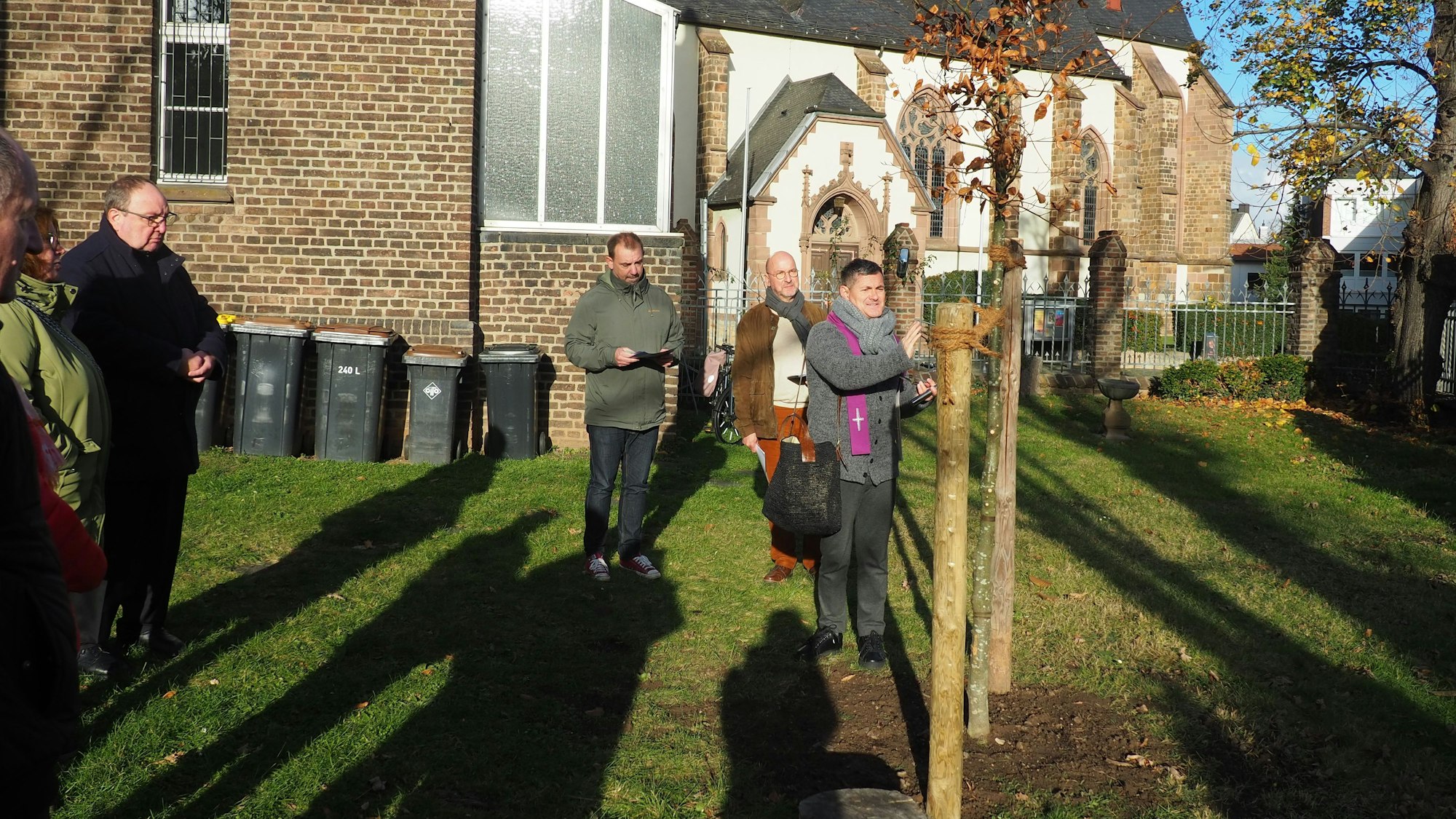Der Geistliche segnet einen Baum, mehrere Menschen schauen zu. Im Hintergrund ist eine Kirche zu sehen.