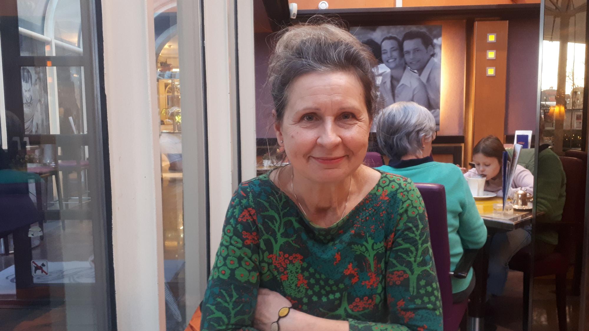 Eine Frau mit grün-rot geblümten Oberteil sitzt lächelnd und mit verschränkten Armen in einem Café.