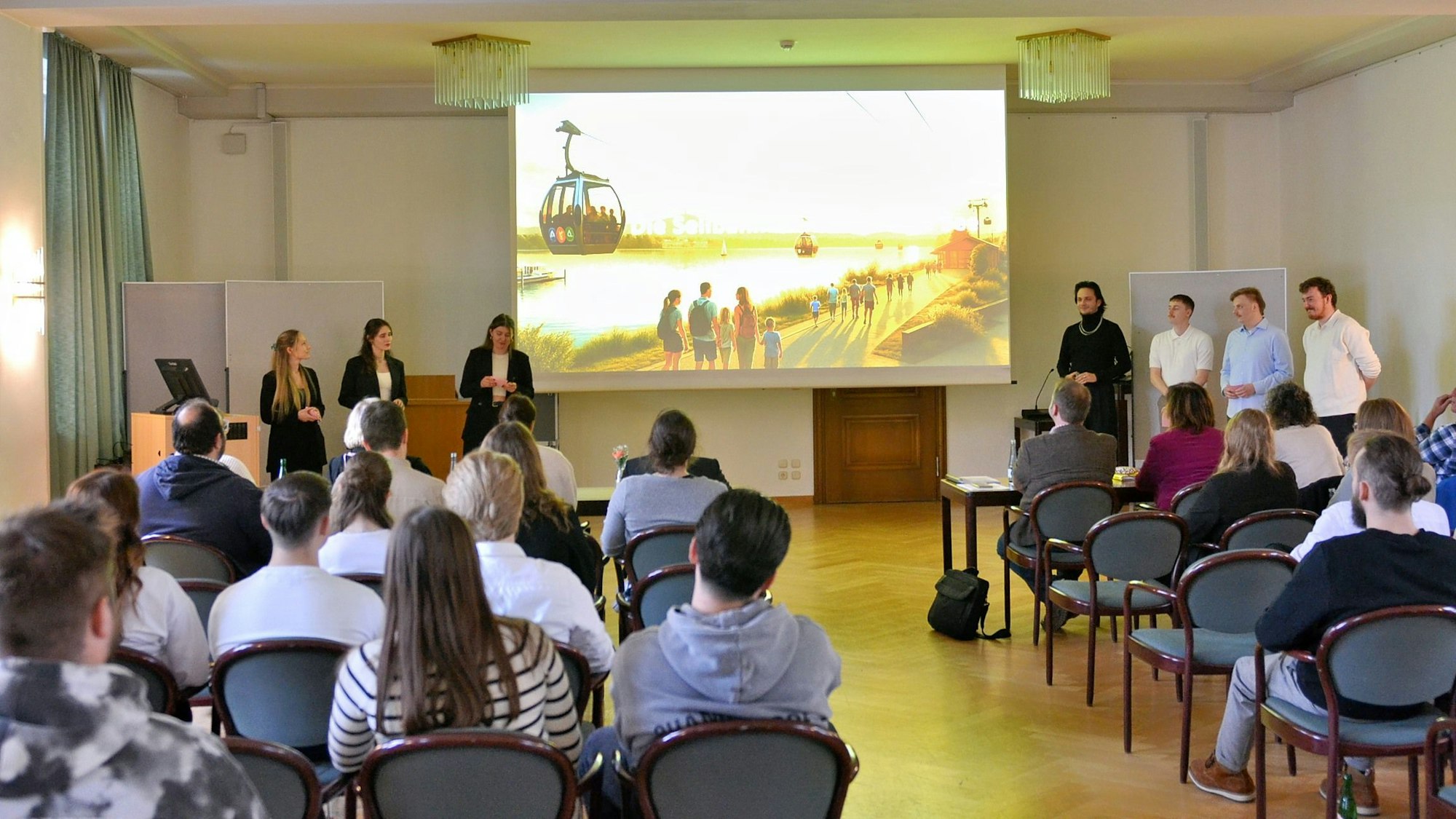 Menschen, von hinten fotografiert, schauen auf eine große Leinwand, die eine Seilbahn zeigt.
