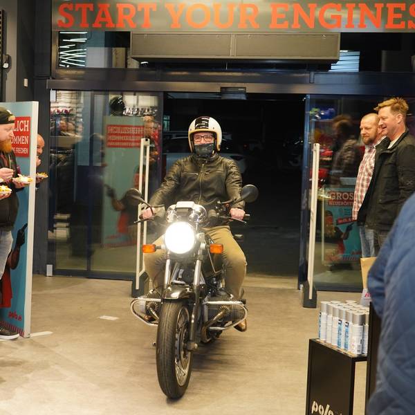 Auf dem Bild ist ein Mann zu sehen, der bei einer Eröffnungsfeier eines Geschäfts mit einem Motorrad in das Ladenlokal fährt.