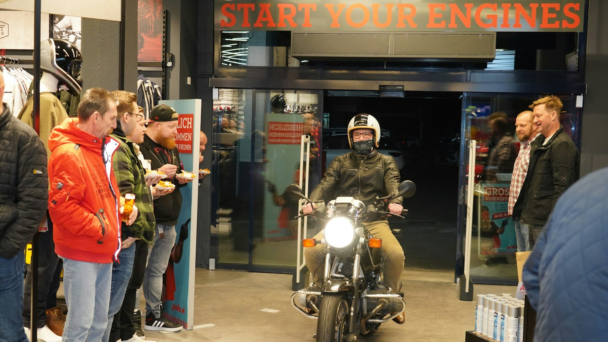 Auf dem Bild ist ein Mann zu sehen, der bei einer Eröffnungsfeier eines Geschäfts mit einem Motorrad in das Ladenlokal fährt.