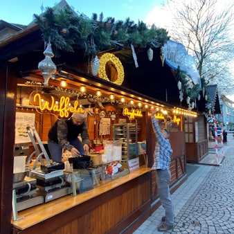 Zu sehen ist einer der Stände des Weihnachtsmarktes. Zwei Mitarbeiter sind mit dem Aufbauen beschäftigt.