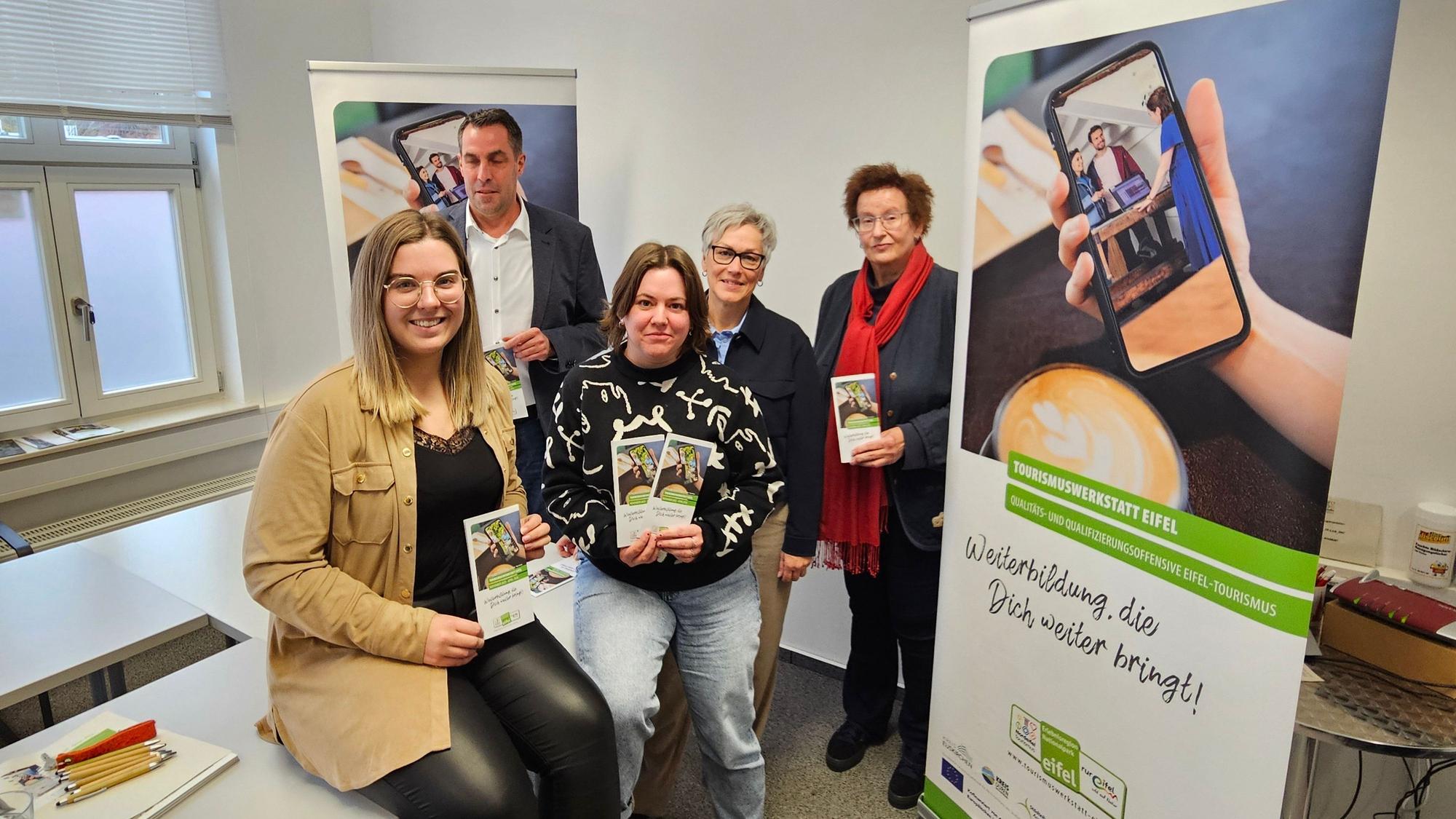 Lara Linden und Verena Koll sitzen auf einem Tisch. Patrick Schmidder, Iris Poth und Barbara Frohnhoff von Rureifel Tourismus stehen dahinter. Alle präsentieren den neuen Flyer.
