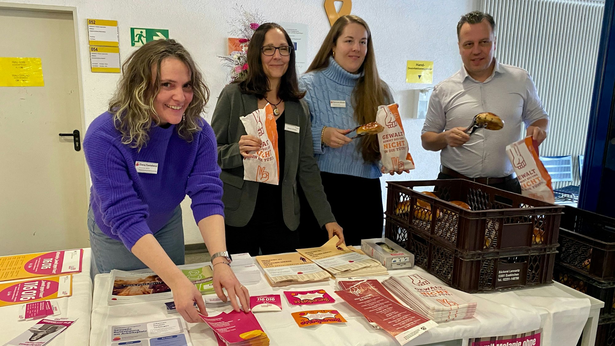 Zwei Mitarbeiterinnen der Frauenberatungsstelle, die Gleichstellungsbeauftragte und der Bürgermeister stehen hinter dem Infostand, an dem es Broschüren und Flyer gibt sowie Weckbrötchen, die in die speziellen Tüten gepackt werden.