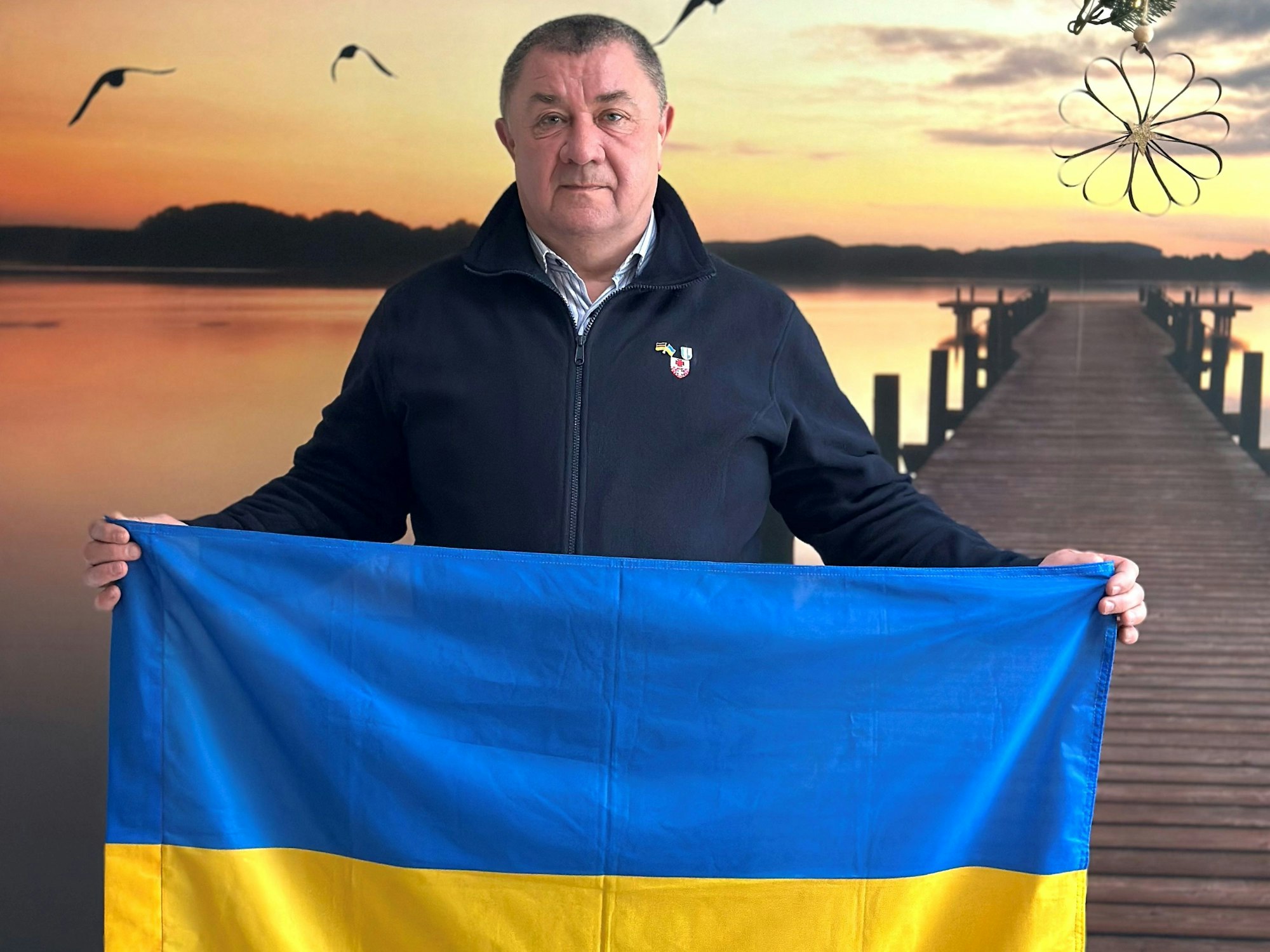 Ein Mann steht vor dem Plakat eines Sonnenuntergangs und hält eine ukrainische Flagge.