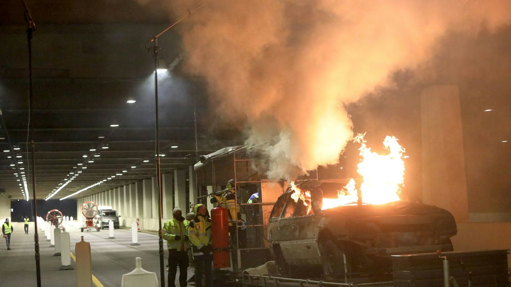 Flammen schlagen aus einem Schrottauto, das sich auf einem Anhänger befindet. Mehrere Personen beobachten das Geschehen.