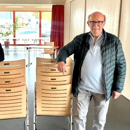 Zwei Männer stehen in einem großen Raum. Sie stützen jeweils einen Arm auf einen Stapel Stühle.