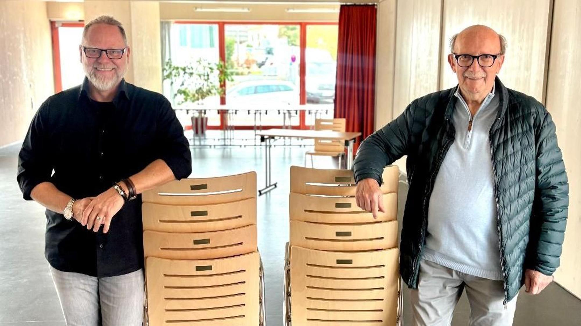 Zwei Männer stehen in einem großen Raum. Sie stützen jeweils einen Arm auf einen Stapel Stühle.