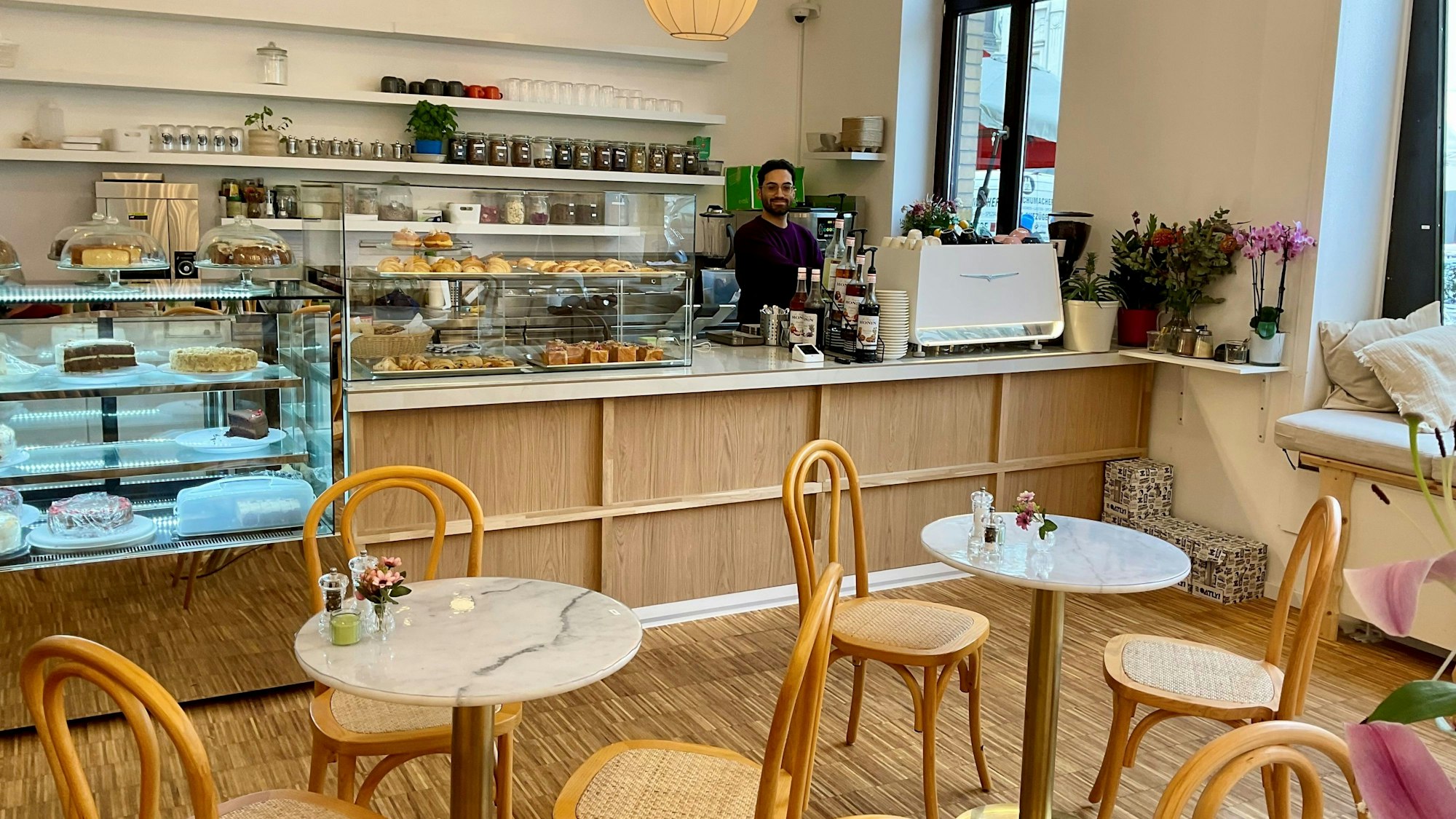 Ein Mann mit dunklem Haar und Brille steht hinter dem Verkaufstresen eines Cafés.