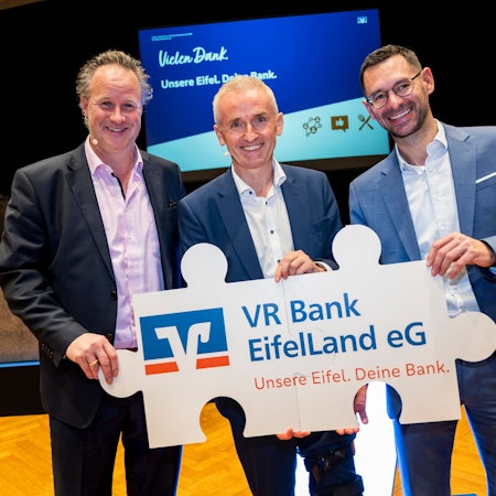 Drei Männer halten ein als Puzzleteile gestaltetes Plakat mit der Aufschrift VR Bank EifelLand eG.