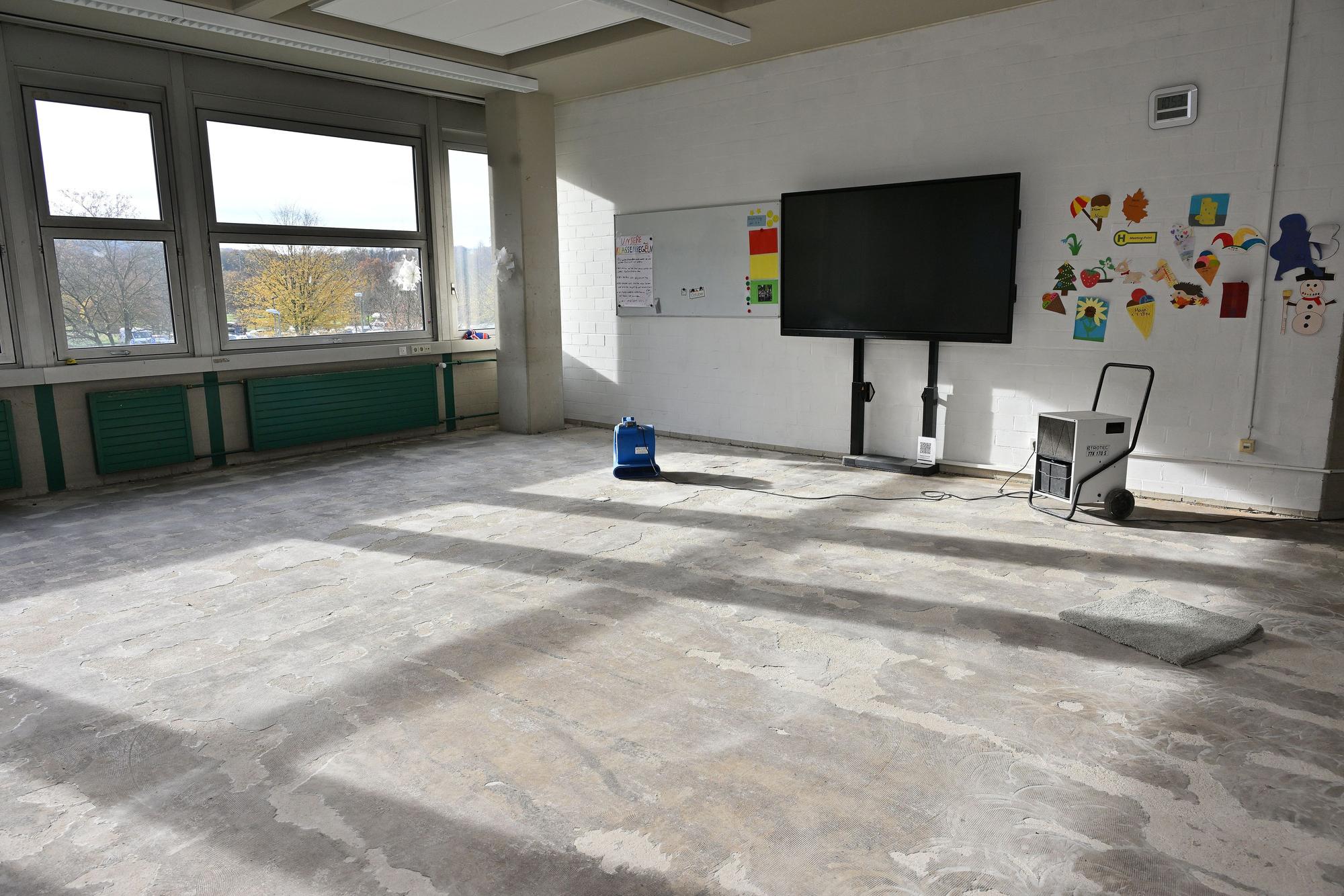 Ein leergeräumtes Klassenzimmer mit feuchtem Estrich und einem Bautrockner darin ist zu sehen.