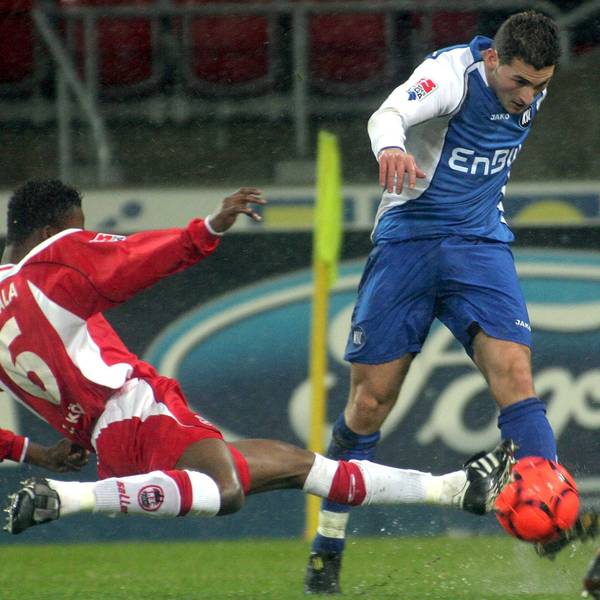 Das Foto zeigt einen Fußballer im blauen Dress, der von seinem Kölner Gegenspieler spektakulär abgegrätscht wird.
