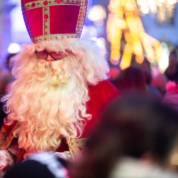 Das Bild zeigt einen Mann mit weißem Bart im Nikolaus-Kostüm. Im Hintergrund funkeln die Lichter des Weihnachtsmarkts.