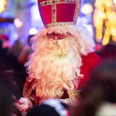Das Bild zeigt einen Mann mit weißem Bart im Nikolaus-Kostüm. Im Hintergrund funkeln die Lichter des Weihnachtsmarkts.