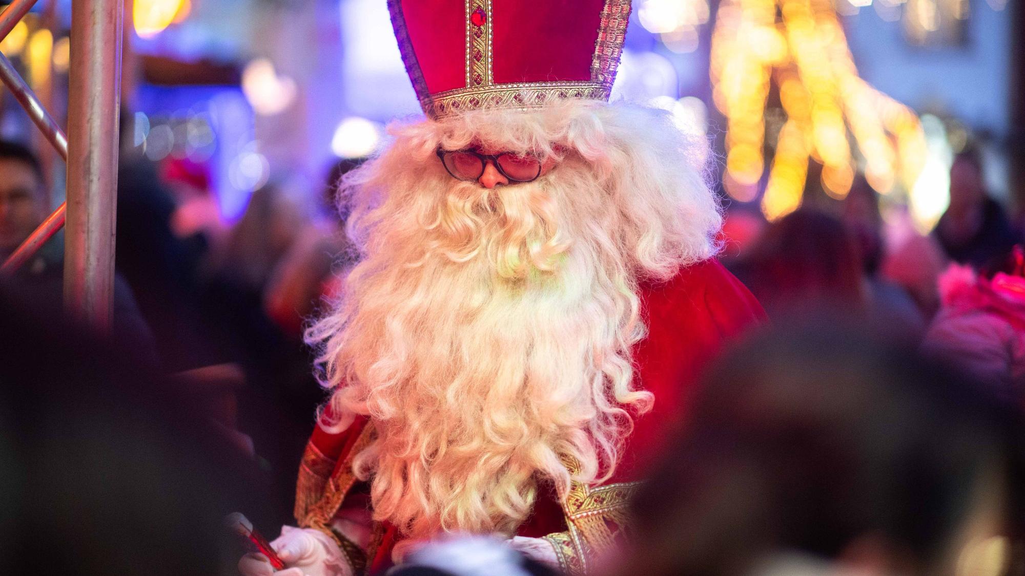 Das Bild zeigt einen Mann mit weißem Bart im Nikolaus-Kostüm. Im Hintergrund funkeln die Lichter des Weihnachtsmarkts.