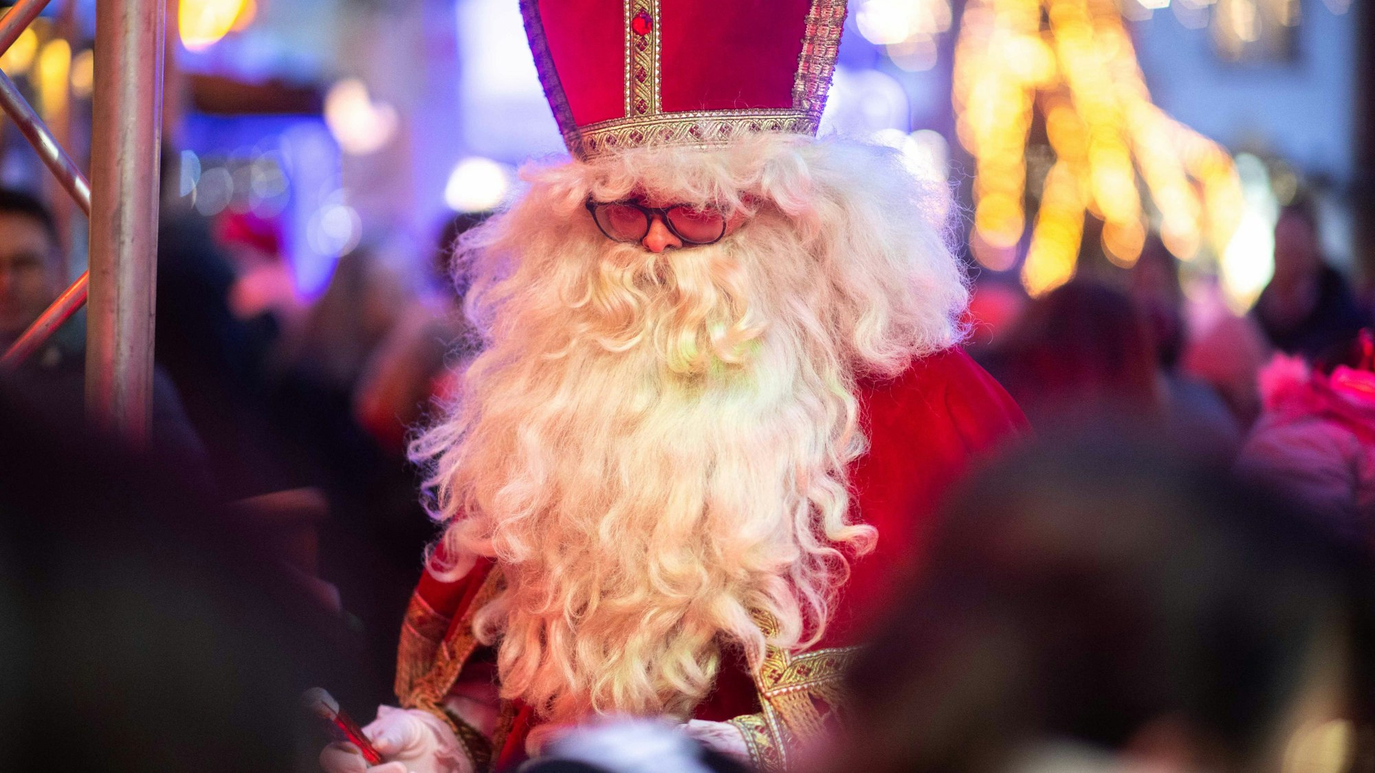 Das Bild zeigt einen Mann mit weißem Bart im Nikolaus-Kostüm. Im Hintergrund funkeln die Lichter des Weihnachtsmarkts.