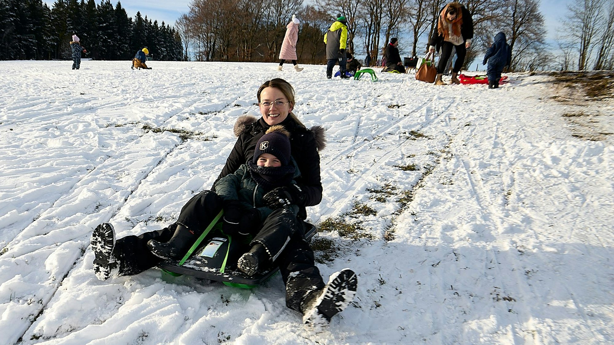 Deborah Schrauben rodelt mit ihrem Sohn den Hang am Weißen Stein hinunter. Im Hintergrund sind weitere Rodler zu sehen. Allerdings lugt aus den Spuren der Kufen wieder Grün durch die geschlossene Schneedecke.