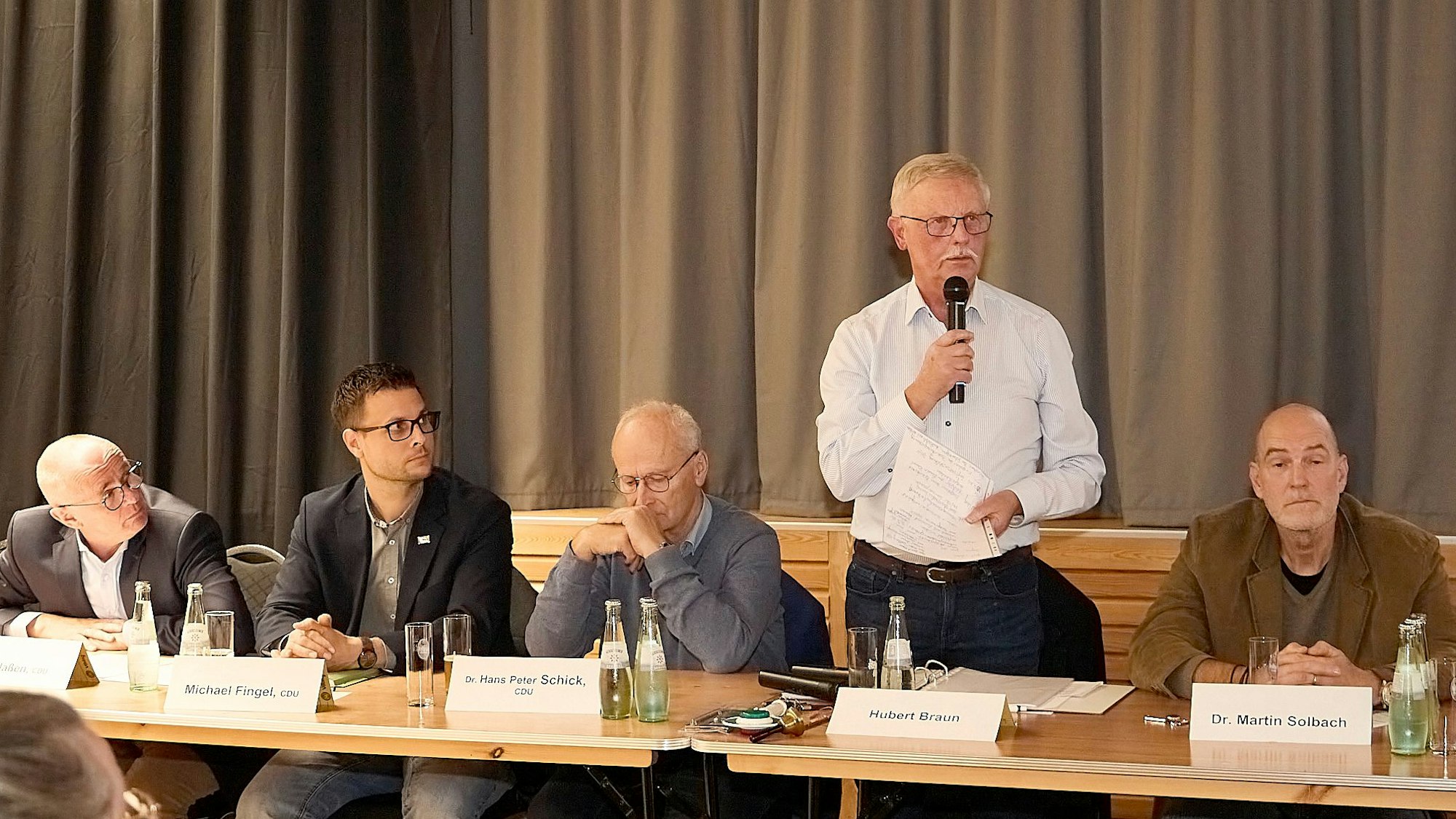 Hubert Braun steht hinter einem Tisch auf dem Podium und hält ein Mikro in der Hand. Neben ihm sitzen unter anderem Mechernichs Bürgermeister Michael Fingel und dessen Vorgänger Dr. Hans-Peter Schick.