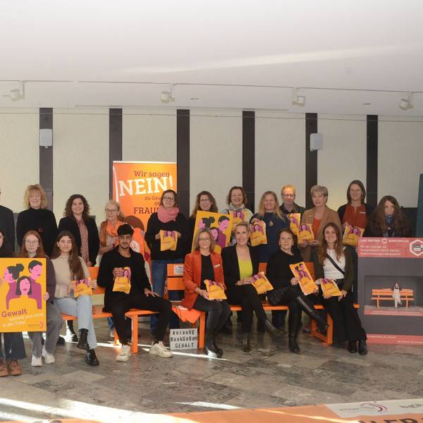 Männer und Frauen sitzen auf orange Bänken. Sie tragen bedruckte Brötchentüten.