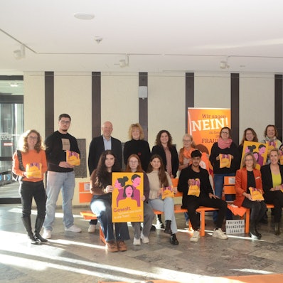 Männer und Frauen sitzen auf orange Bänken. Sie tragen bedruckte Brötchentüten.