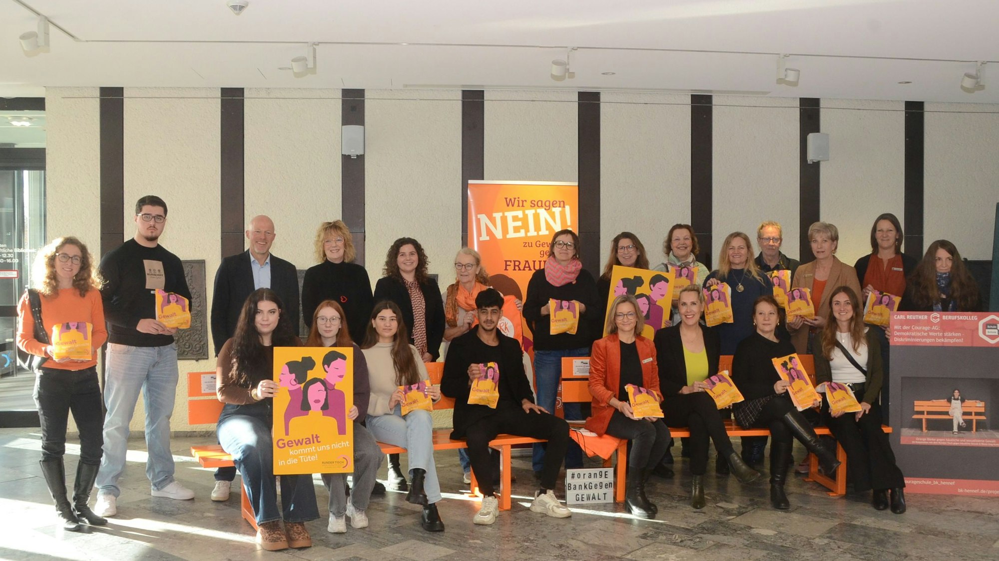 Männer und Frauen sitzen auf orange Bänken. Sie tragen bedruckte Brötchentüten.