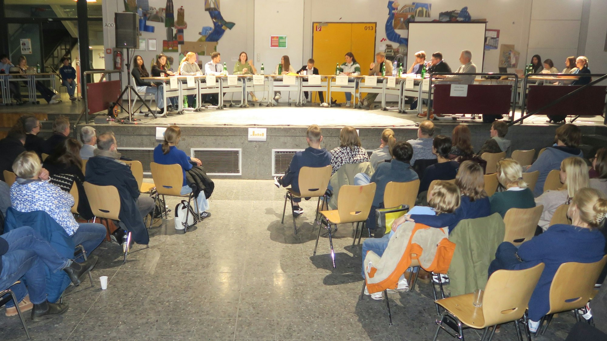 In einer Schulaula sitzen Menschen im Publikum, auf der Bühne ist ein halbkreisförmiges Panel mit vielen Teilnehmern zu sehen.