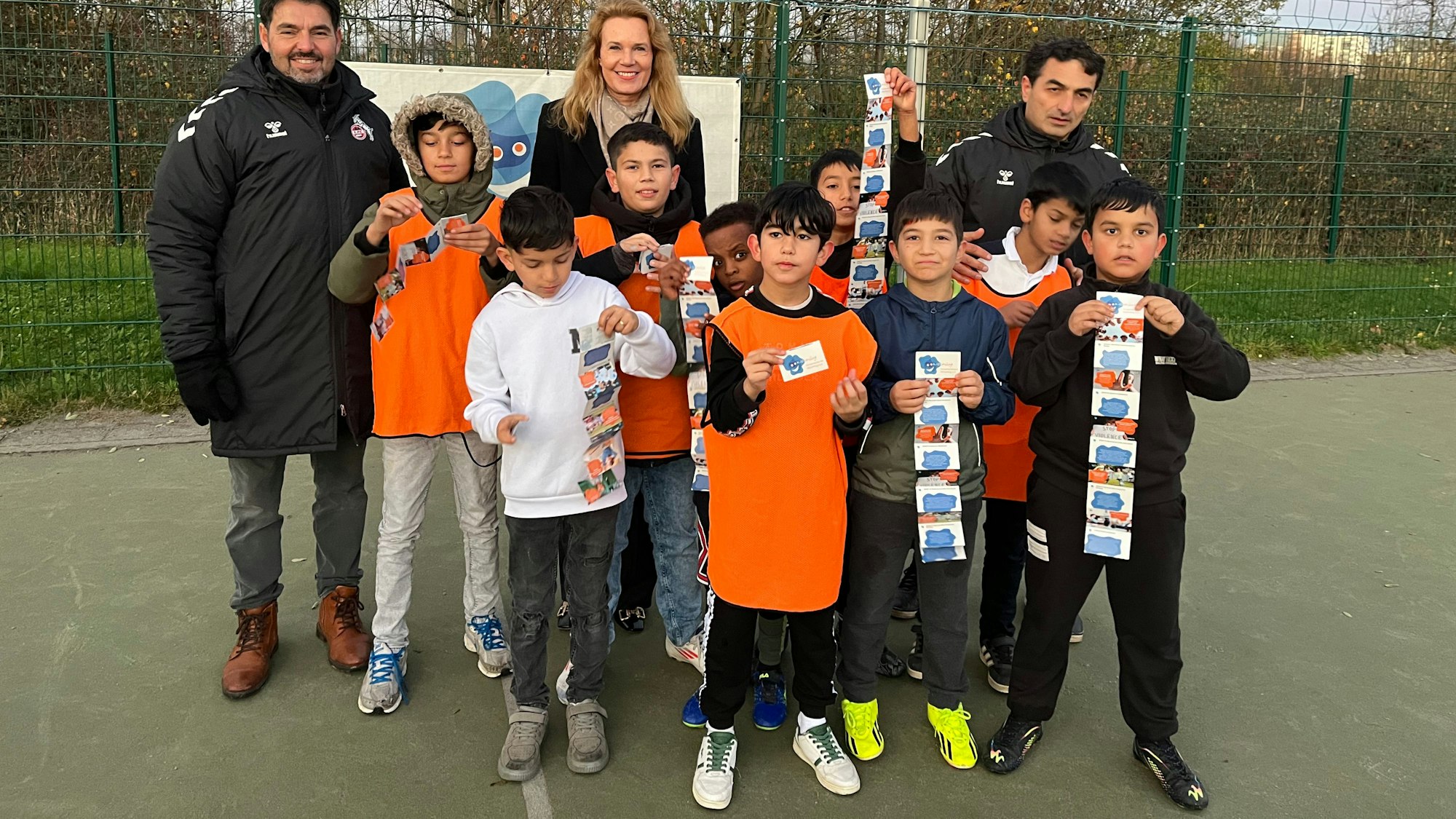 Zwei Männer in Trainingsjacken, eine blonde Frau und viele Kinder stehen auf einem Trainingsplatz zusammen.