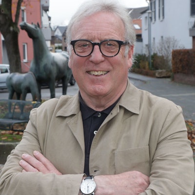Ein Mann mit weißen Haaren und einer Brille mit schwarzem Gestell lächelt in die Kamera.