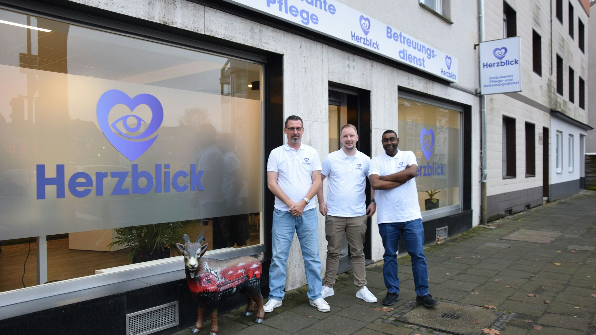 Drei Männer stehen neben der Figur von Hennes, dem Geißbock, vor der Zentrale ihres neuen Pflegedienstes Herzblick.