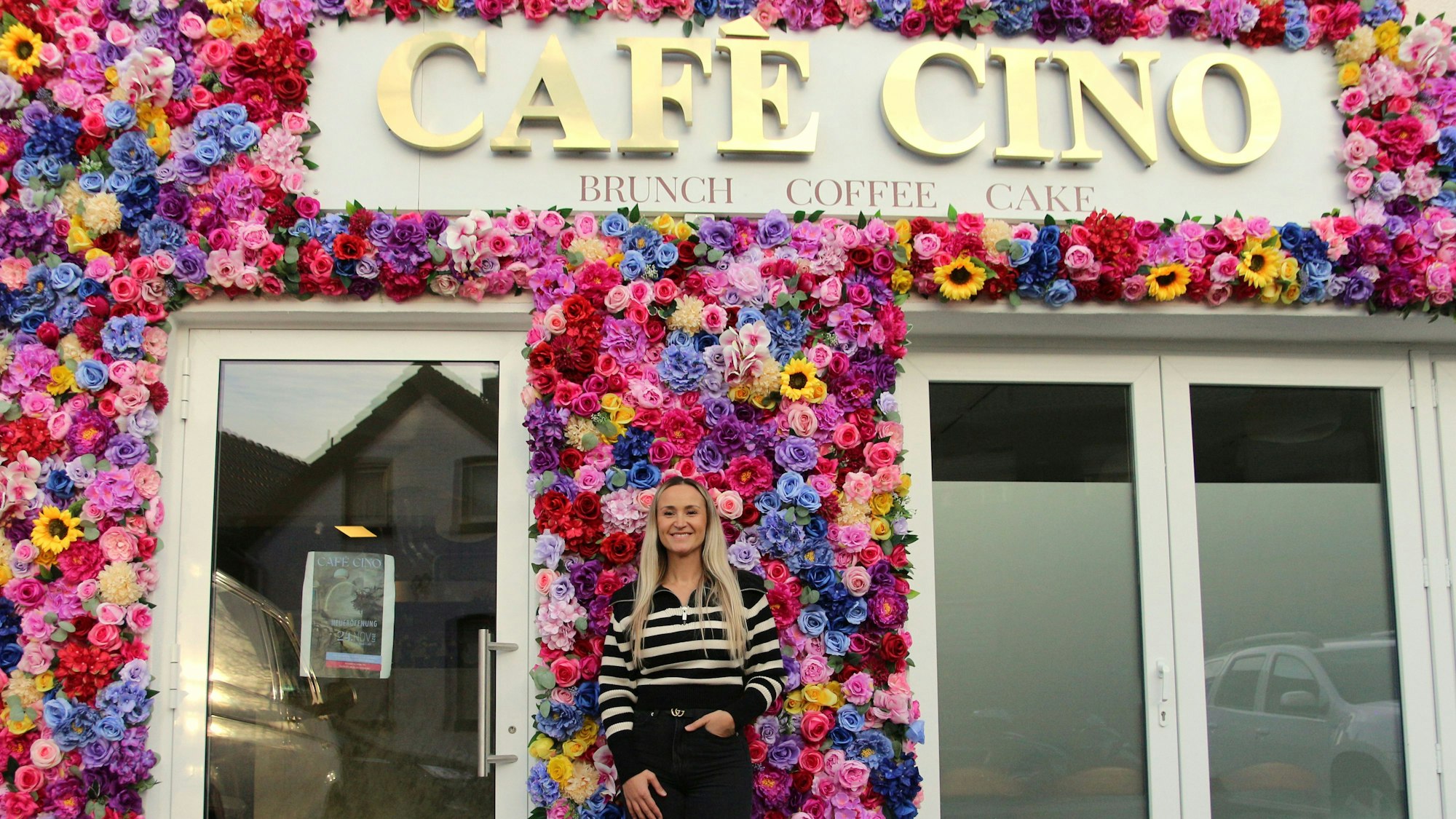 Eine Frau steht vor einer Fassade, die mit vielen Blumen geschmückt ist und den Schriftzug „Café Cino“ trägt.