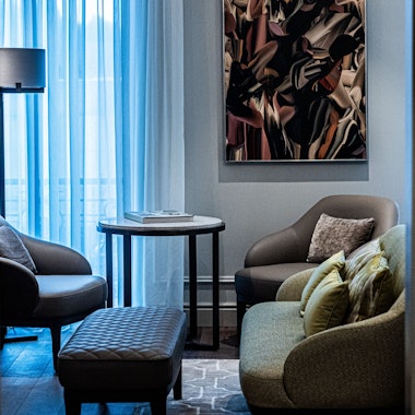 Der Blick durch die Tür der Suite zeigt ein kleines Sofa, vor der eine Lederbank steht. Im Hintergrund sind zwei Lederstühle mit einem Beistelltisch vor dem Fenster zu sehen.