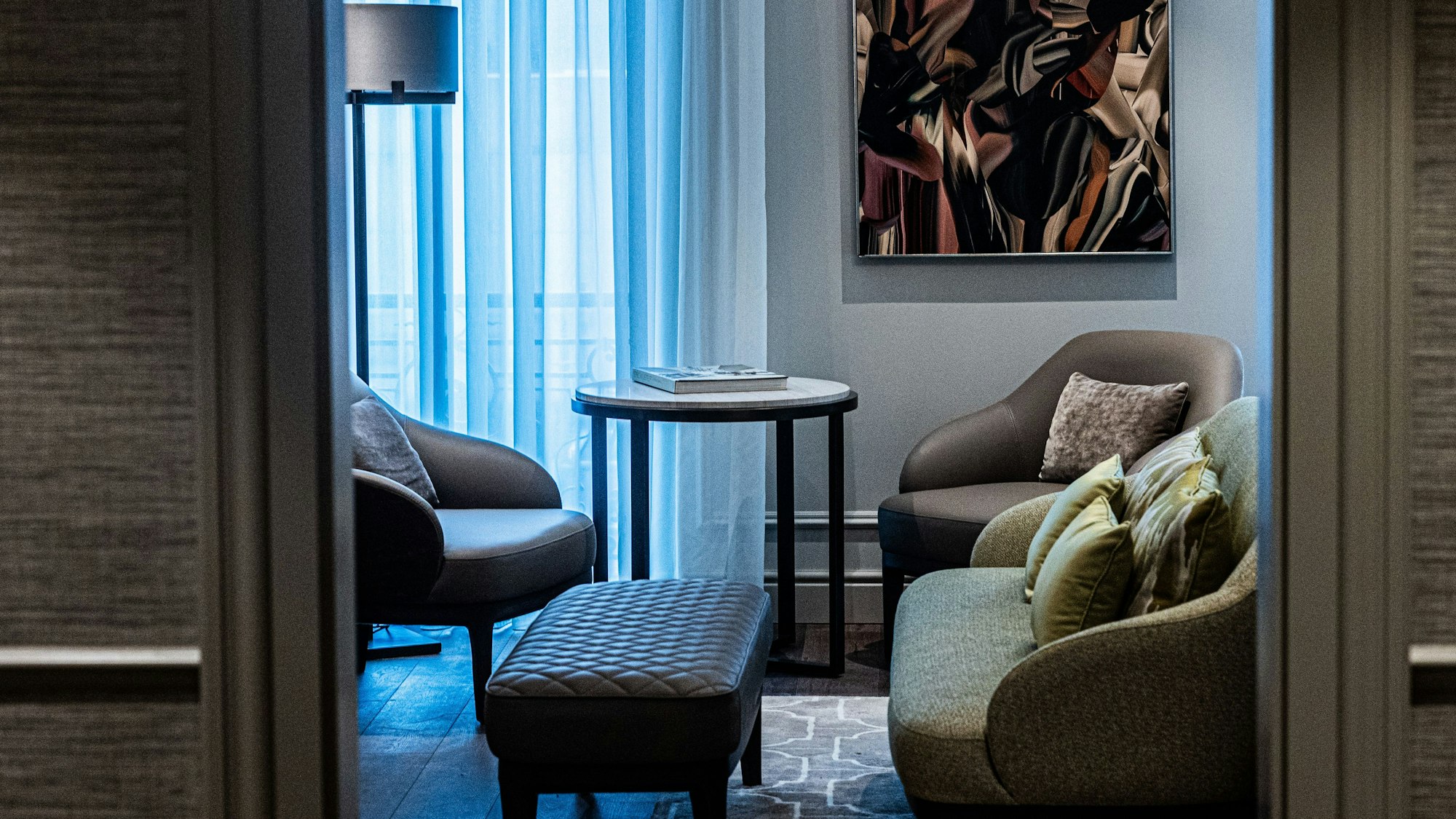 Der Blick durch die Tür der Suite zeigt ein kleines Sofa, vor der eine Lederbank steht. Im Hintergrund sind zwei Lederstühle mit einem Beistelltisch vor dem Fenster zu sehen.