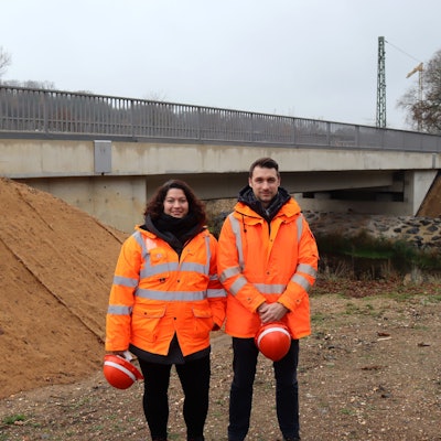 Ein Mann und eine Frau in orangefarbener Jacke stehen vor einer Brücke. Beide halten einen Helm in ihren Händen.