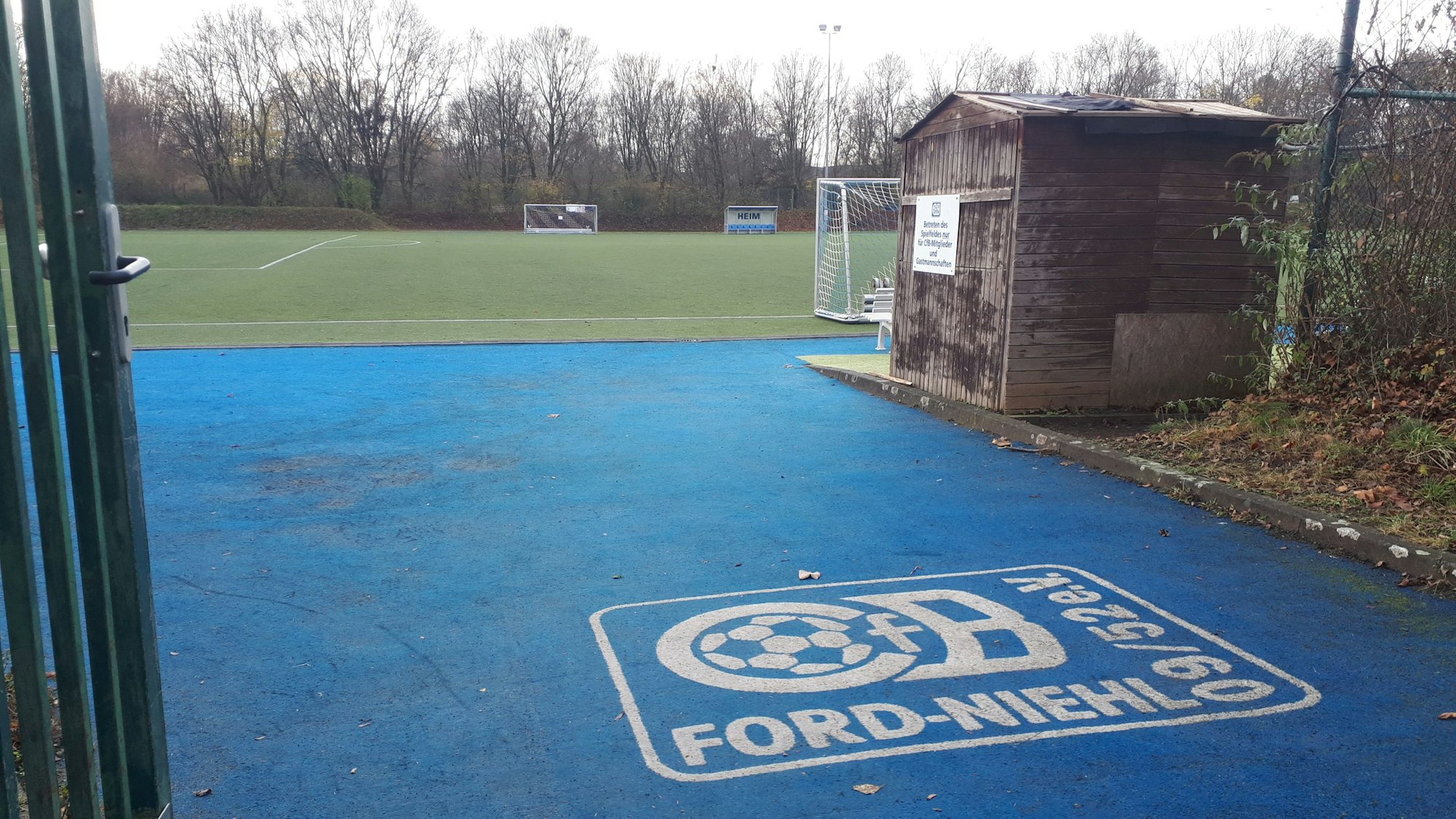 Ein blauer Bodenbelag hinter einem geöffneten Gittertor führt zu einem Trainingsgelände.