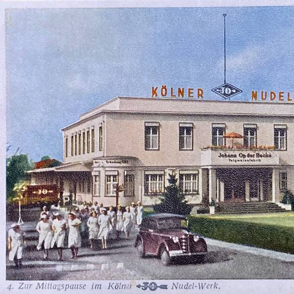 Frauen in weißen Kleidern und ein Auto stehen vor einem stattlichen Gebäude, auf dem der Schriftzug „Kölner JO Nudeln“ steht.