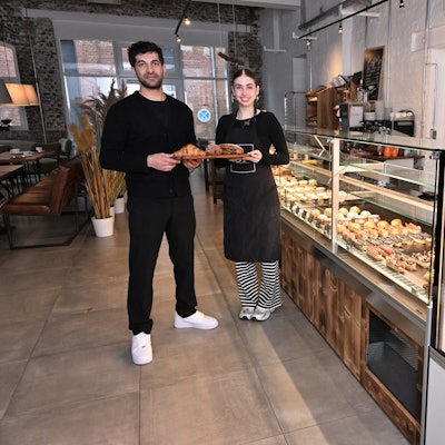 Die beiden stehen mit einem Tablett mit Gebäck vor der Theke.