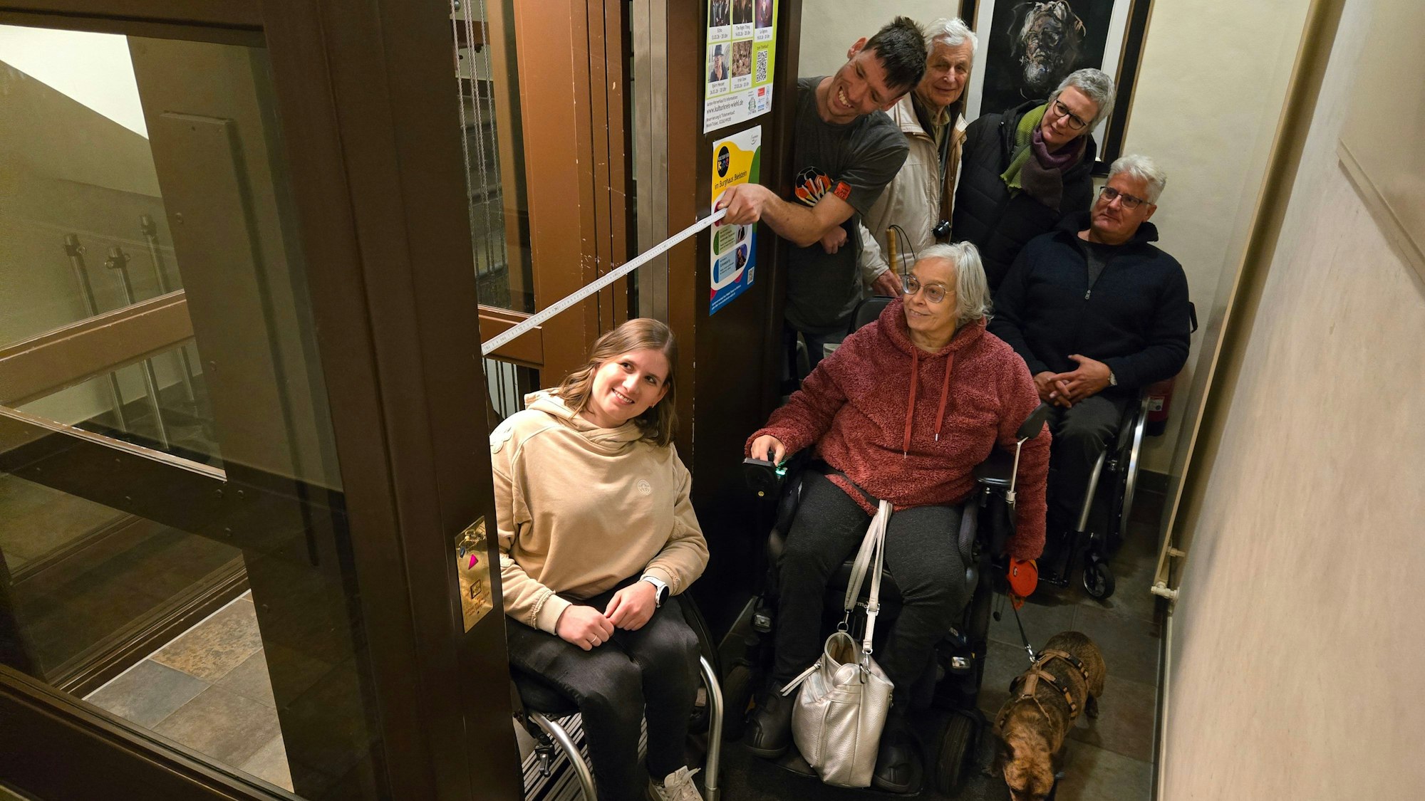 Eine Gruppe von Menschen, einige im Rollstuhl, einer misst die Breite eines Fahrstuhlzugangs.
