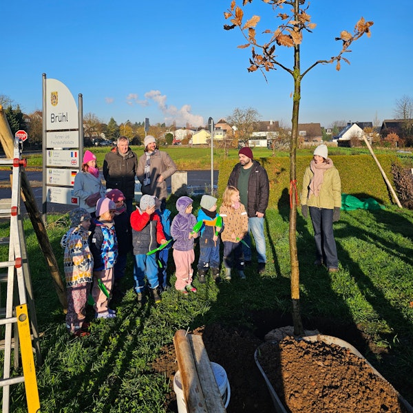 Kinder und Erwachsene stehen neben einem neu gepflanzten Baum.